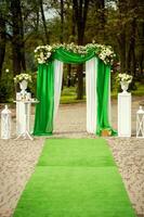 Bogen zum das Hochzeit Zeremonie, dekoriert mit Grün und Weiß Stoff und Blumen draußen. lange Grün Teppich zu das Hochzeit Bogen auf das Wald Hintergrund. foto
