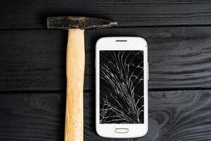 Smartphone zerschlagen durch Hammer isoliert auf schwarz hölzern Hintergrund. gebrochen Berührungssensitiver Bildschirm von ein Smartphone Lügen schließen zu das Hammer auf Tisch. foto
