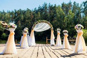 schön Platz zum Hochzeit Zeremonie auf das hölzern Brücke Über das Fluss. Torbogen dekoriert mit cremig und Weiß Stoff und Blumen auf das natürlich Wald Hintergrund im Sommer. foto