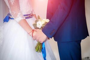 schön jung Paar Stehen schließen zu einer Ein weiterer während das Hochzeit Zeremonie. Braut im Weiß Kleid mit Blau Gürtel und Bräutigam tragen elegant passen halten Hände zusammen. foto