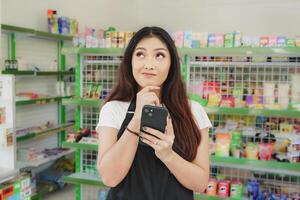 nachdenklich asiatisch Menschen halten ihr Telefon, Kassierer ist tragen schwarz Schürze Stehen im ein Lebensmittel foto