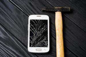 Berührungssensitiver Bildschirm von ein Smartphone ist gebrochen mit ein Hammer auf das grau hölzern Hintergrund. beschädigt Handy, Mobiltelefon Telefon mit Hammer auf Tisch. foto