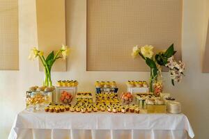 Süßigkeiten Bar. ein festlich Tabelle mit Panakota, Makronen, Muffins, Baiser und mit zwei Vasen mit schön Blumen ist gelegen in der Nähe von das Mauer beim das Hochzeit im das Restaurant foto