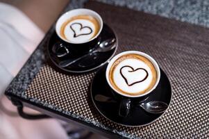 zwei schwarz Tassen auf Untertassen mit Cappuccino auf ein klein Tabelle im das Zimmer. Herz Symbole ist gemalt auf das Weiß Oberfläche von Kaffee mit Milch. Nahansicht foto