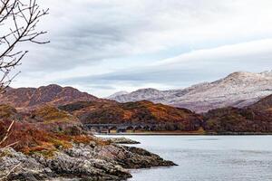 heiter Landschaft mit ein still See, herbstlich Bäume, und entfernt schneebedeckt Berge unter ein wolkig Himmel im Schottland. foto