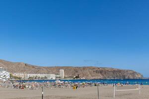 sonnig Strand mit Touristen und klar Blau Himmel, Berg Hintergrund, und Küsten Gebäude im los Cristianos, Teneriffa. foto