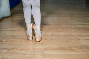 Beine von ein wenig Ballerina im ein Gold Ballett Schuhe und Weiß Socken posieren auf Zehenspitzen im Ballett Halle auf das Boden. Nahaufnahme. foto