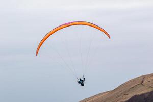 Gleitschirm im Flug mit Orange Flügel gegen ein klar Himmel Über ein Berg Steigung im Gipfel Bezirk, England. foto