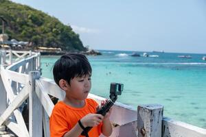 ein wenig asiatisch Junge ist filmen ein online Video draußen während gehen zu das Strand. foto