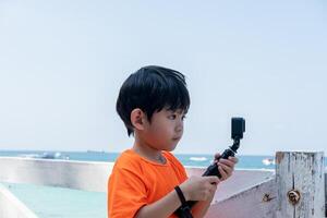 ein wenig asiatisch Junge ist filmen ein online Video draußen während gehen zu das Strand. foto