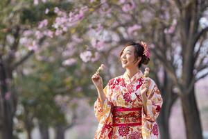 japanisch Frau im traditionell Kimono Kleid halten Süss Hanami Dango Dessert während Gehen Innerhalb Park beim Kirsche blühen Baum während Frühling Sakura Festival foto