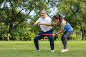Senior asiatisch Mann ist mit Gummi Band zu bauen oben seine Bein Muskel Stärke während seine Tochter ist Jubel oben im das Öffentlichkeit Park zum ältere Langlebigkeit Übung und draussen trainieren foto