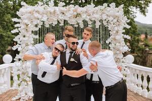 heiter, jung, energisch Zeugen von das Bräutigam Nächster zu das Bräutigam. freunde gratulieren das Bräutigam. das Bräutigam im ein Weste und seine freunde im Weiß Hemden Pose in der Nähe von das Hochzeit Bogen. Hochzeit im Natur. foto