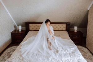 schön Braut im Weiß Hochzeit Kleid Sitzung auf das Bett im das Morgen. Mode Schuss, schön Frau im ein Dressing Kleid. Mode, Glanz Konzept foto