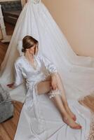 ein schön Braut ist Sitzung im ein Dressing Kleid im das Morgen Vor das Hochzeit Zeremonie im ein Hotel mit ein modern Innere. unglaublich Frisur von das Braut. natürlich und modern bilden. foto