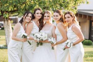 ein Brünette Braut und ihr Brautjungfern im identisch Weiß Kleider sind Stehen auf das Hintergrund von Natur mit Blumensträuße von Blumen im ihr Hände. Mädchen im identisch Kleider sind Herstellung aus beim ein Hochzeit foto