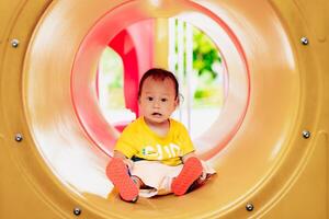Porträt von Kind spielen auf Spielplatz Konzept.süß asiatisch Baby Junge ist Sitzung im Gelb Orange Rohre, auf ein heiß Tag, Sommer- oder Frühling Jahreszeit, Kleinkind Kind einer Jahr drei Monate alt. echt Personen. foto