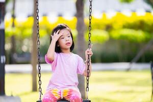 asiatisch wenig Kind Mädchen auf schwingen beim das Park, gefüllt mit Freude und Lachen, schwingen glücklich im das Sommer- Sonne, umgeben durch Natur und spielerisch Spaß, Kinder alt 6 Jahre alt, Sommer- oder Frühling mal. foto