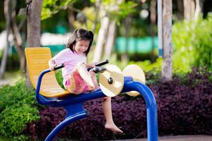 asiatisch Kind Mädchen genießen ein sonnig Tag im das Park, Sitzung auf Schaukeln und ein Bank, oder Bein Übung Maschine, umgeben durch Natur und lächelt im das Park, glücklich Lächeln Kind aktiv im Sommer- oder Frühling mal foto