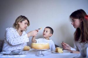 Familie von drei, Mutter, Teenager Tochter und wenig Sohn, Essen Kuchen im Pyjama beim ein Tabelle mit Tulpen foto
