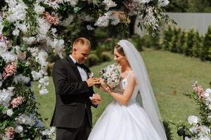das Bräutigam setzt auf das Braut Hochzeit Ring während das Hochzeit Zeremonie in der Nähe von das Blume Bogen. Sommer- Hochzeit im Natur. sie genannt Ja. ein berühren Moment beim das Hochzeit Zeremonie foto