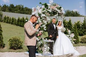 Hochzeit Zeremonie im Natur. das Braut und Bräutigam in der Nähe von das Blume Bogen. das Meister von Zeremonien beim das Hochzeit während das Performance im das Hintergrund von das Braut und Bräutigam. foto