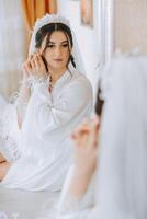 Schön, jung, perfekt Modell- Braut im ein Weiß Spitze Kleid sitzt im das Zimmer, Schlafzimmer im das Morgen, Putten ein Ohrring auf ihr Ohr. Hochzeit Fotografie, Porträt, Konzept. foto