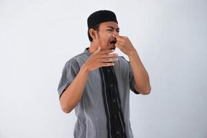 Porträt von angeekelt jung Muslim asiatisch Mann kneift Nase mit Finger Hände sieht aus mit der Ekel etwas stinkt Schlecht Geruch Situation tragen Muslim Kleider isoliert auf Weiß Hintergrund foto