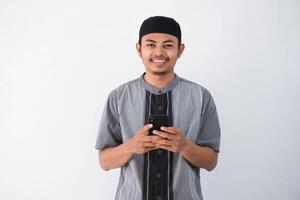glücklich oder lächelnd jung asiatisch Muslim Mann halten Telefon zellular suchen Kamera tragen koko Kleider isoliert auf Weiß Hintergrund. Ramadan Konzept foto