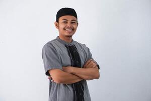 Lächeln oder glücklich jung asiatisch Muslim Mann gekreuzt Waffen und suchen Kamera tragen grau koko Kleider isoliert auf Weiß Hintergrund foto