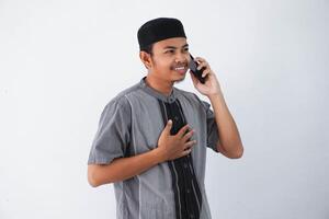 lächelnd jung asiatisch Muslim Mann halten Truhe und halten Telefon Berufung durch Handy, Mobiltelefon Telefon tragen koko Kleider isoliert auf Weiß Hintergrund foto