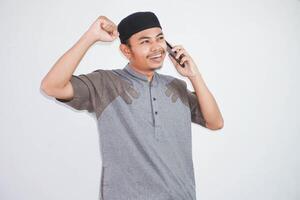 aufgeregt oder glücklich jung asiatisch Muslim Mann reden auf Handy, Mobiltelefon Telefon und tun Gewinner Geste tragen koko Kleider isoliert Über Weiß Hintergrund foto