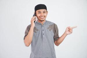 heiter jung asiatisch Muslim Mann reden auf Handy, Mobiltelefon Telefon und zeigen Finger Weg beim Kopieren Raum tragen grau Muslim Kleider isoliert auf Weiß Hintergrund foto