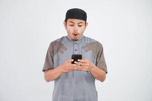schockiert jung asiatisch Muslim Mann halten Handy, Mobiltelefon Telefon tragen Muslim Kleider isoliert auf Weiß Hintergrund foto