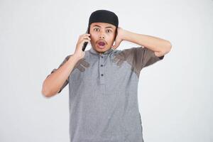 schockiert jung asiatisch Muslim Mann reden auf Handy, Mobiltelefon Telefon Stecker Ihre Ohren tragen grau Muslim Kleider isoliert auf Weiß Hintergrund foto