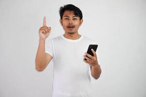 aufgeregt jung asiatisch Mann zeigen Finger oben haben ein gut Idee mit halten Handy, Mobiltelefon Telefon tragen Weiß t Hemd isoliert auf Weiß Hintergrund foto