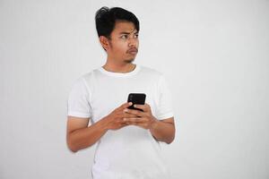 nachdenklich jung asiatisch Mann mit ein ernst Gesicht Denken Über ein Frage, halten Handy, Mobiltelefon Telefon tragen Weiß t Hemd isoliert auf Weiß Hintergrund foto