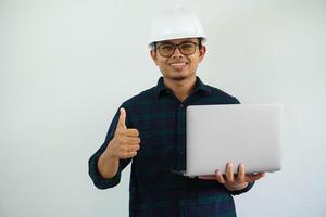jung asiatisch Architekt Mann tragen Baumeister Sicherheit Helm Über isoliert Hintergrund tun glücklich Daumen oben Geste und halten Laptop mit Hand. foto