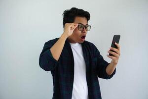 jung asiatisch Mann suchen beim seine Telefon mit ein überrascht Ausdruck während halten Brille im seine Hände isoliert auf Weiß Hintergrund foto