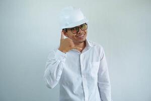 jung asiatisch Mann ist Ingenieur tragen Helm Stehen mit zeigen ein Handy, Mobiltelefon Telefon Anruf Geste mit Finger und lächelnd mit zuversichtlich, Architekt oder Auftragnehmer, Arbeiter oder Arbeit, industriell Konzept. foto