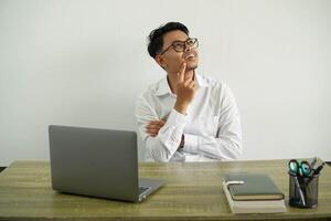 jung asiatisch Geschäftsmann im ein Arbeitsplatz Denken ein Idee während suchen hoch, tragen Weiß Hemd mit Brille isoliert foto