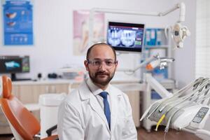 zuversichtlich Zahnarzt im Stomatologie Kabinett mit Orange Ausrüstung tragen Dental Uniform. medizinisch Spezialist im Oral Hygiene tragen Labor Mantel suchen beim Kamera im Zahnheilkunde Büro. foto