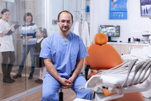 Fachmann Zahnarzt tragen Schutz und geduldig diskutieren mit Kieferorthopäde. Brille stomatologe im beruflich Zähne Klinik lächelnd tragen Uniform suchen beim Kamera. foto