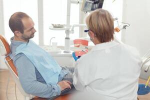 Stomatologe halten sanitär Modell- von das Unterkiefer reden mit krank Mann. Senior Zahnarzt erklären richtig Dental Hygiene mit Attrappe, Lehrmodell, Simulation Skelett von Zähne während Arbeiten im Stomatologie Krankenhaus foto