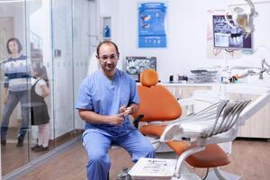 Fachmann Zahnarzt diskutieren Über mehrere Zähne Hygiene Probleme Sitzung auf Stuhl. stomatologe im beruflich Zähne Klinik lächelnd tragen Uniform suchen beim Kamera. foto