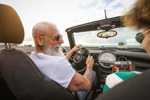 glücklich Senior Paar haben Spaß nehmen Foto mit alt Jahrgang Kamera während Fahren auf Neu Cabrio Auto während Straße Ausflug Tour Ferien