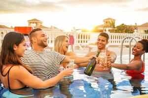 Gruppe von glücklich freunde Herstellung ein Schwimmbad Party Toasten mit Flasche von Champagner beim Sonnenuntergang - - jung Menschen haben Spaß Trinken mit funkelnd Wein im Luxus tropisch Resort - - Jugend Lebensstil Konzept foto