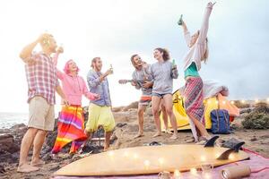 glücklich freunde haben Spaß spielen Gitarre und Tanzen auf das Strand - - Gruppe von jung Menschen Herstellung Party Trinken Biere während Camping mit Zelt - - reisen, Ferien und Jugend Ferien Lebensstil Konzept foto