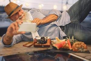 Senior Mann lächelnd während nehmen Selfie mit Handy, Mobiltelefon Smartphone Kamera - - glücklich Rentner haben Spaß auf Boot Party Trinken Champagner Essen tropisch Früchte - - Alten Menschen und Technologie Konzept foto