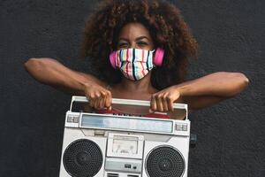 jung afro Frau tragen Gesicht Maske Tanzen draussen während Hören zu Musik- mit kabellos Kopfhörer und Jahrgang Boombox Stereo foto